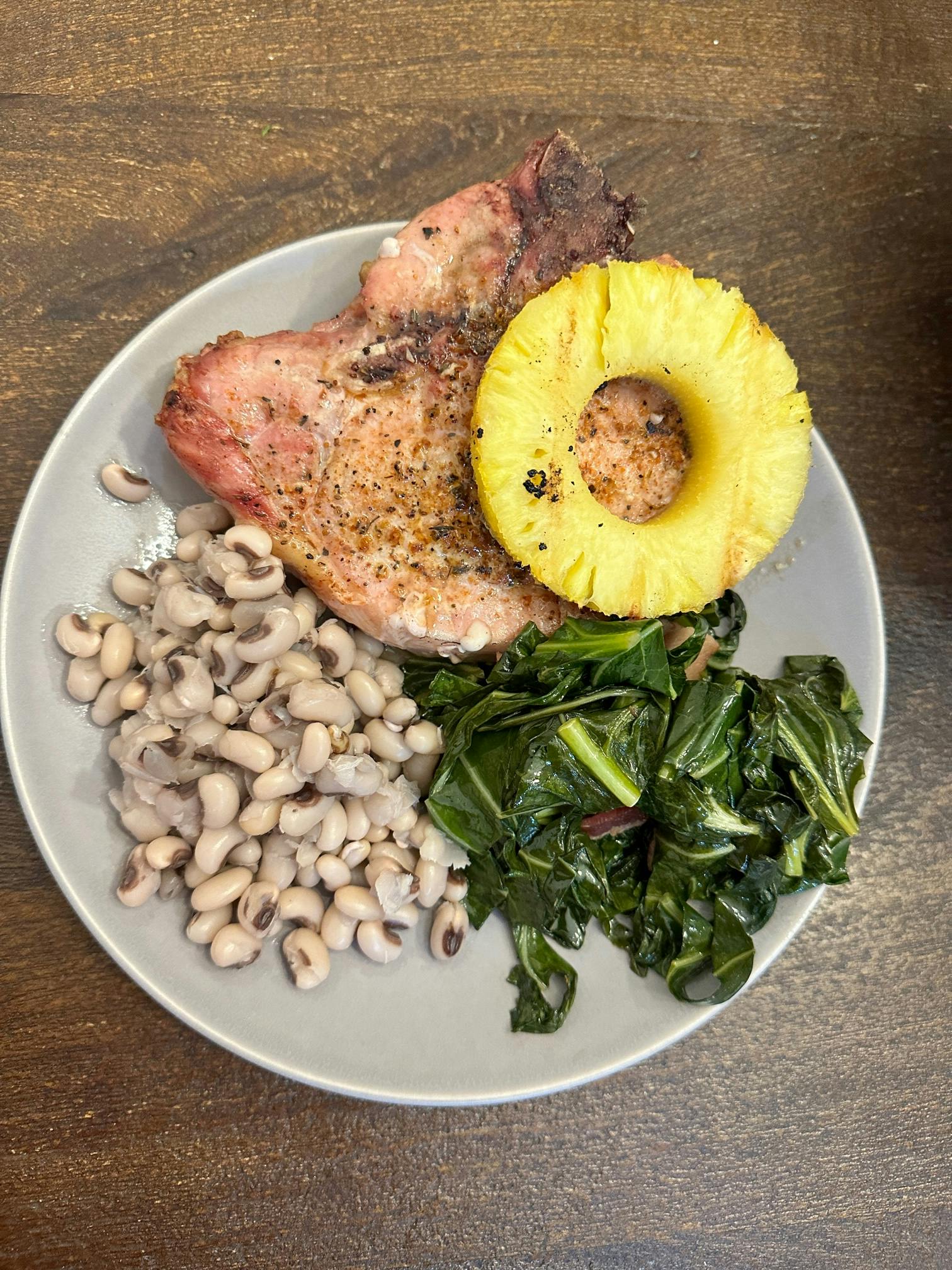 A plate with black eyed peas, collard greens and a pork chop with a piece of pineapple on top of it